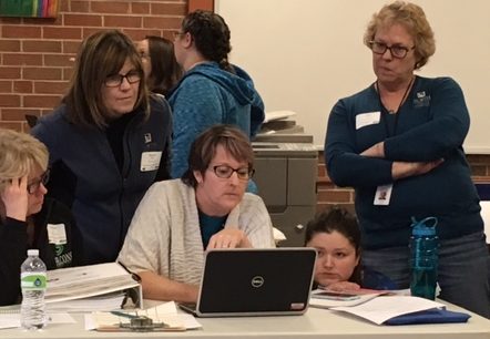 Woman at desk with laptop and students standing around, looking at laptop and learning lessons.